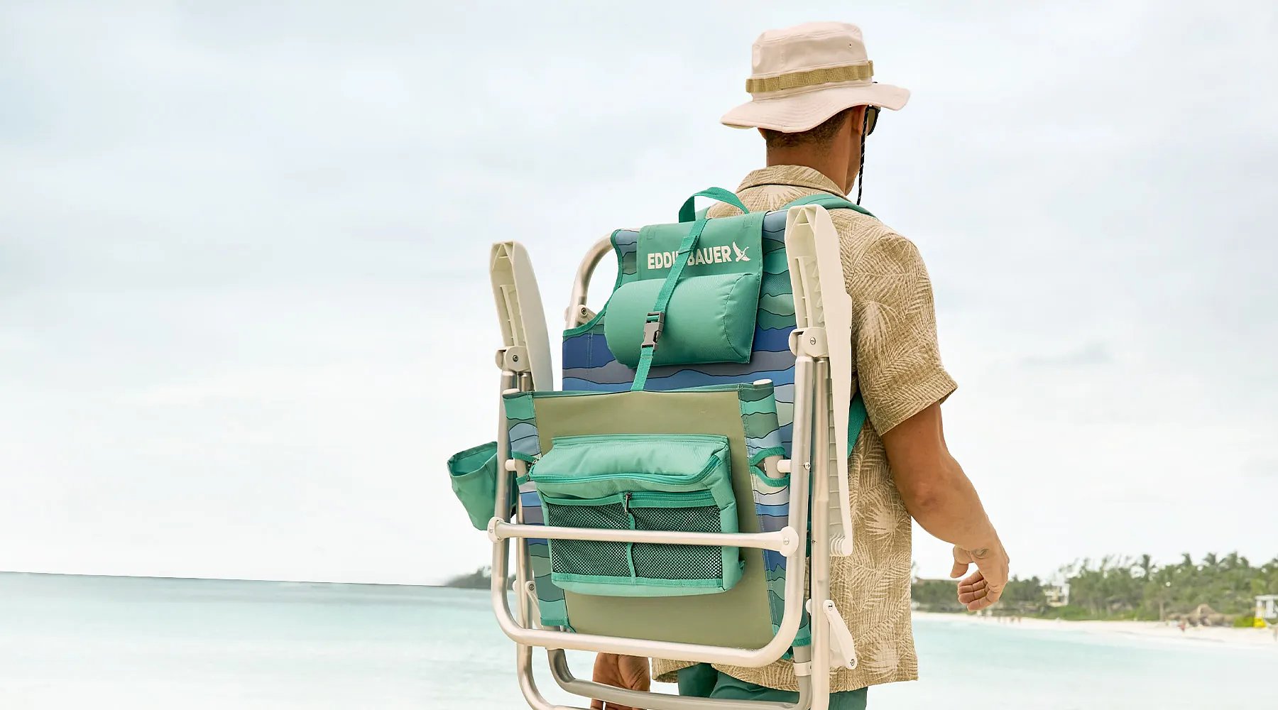 A man carrying a backpack chair walks on a tropical beach.