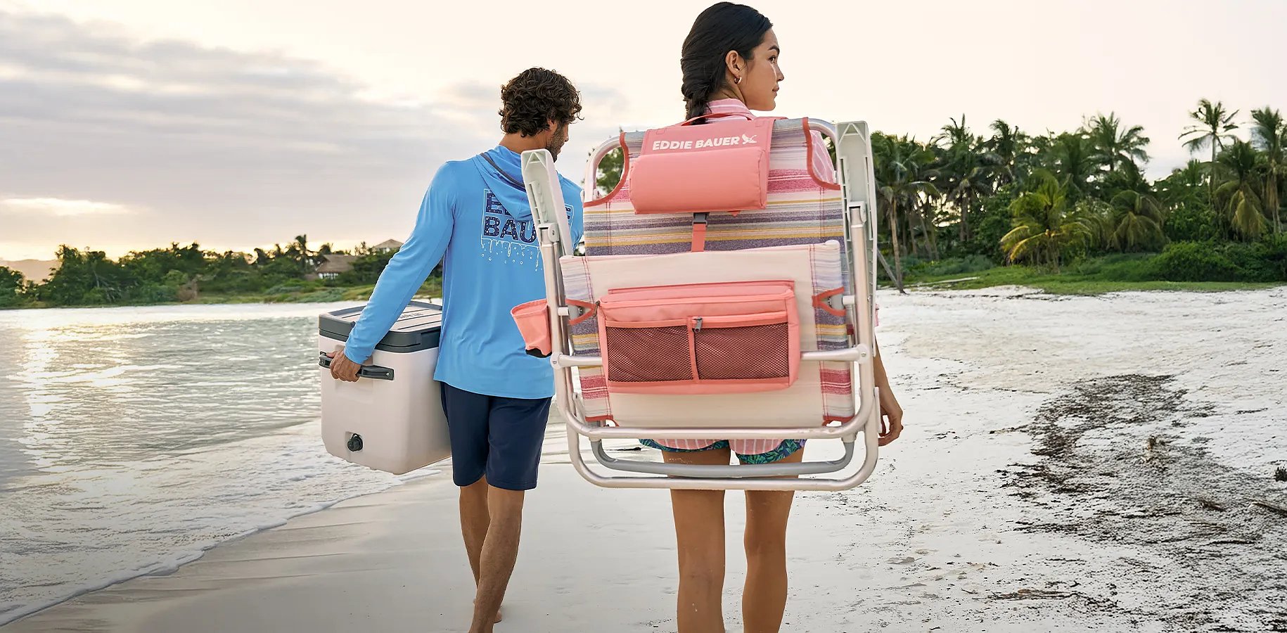 A woman carrying a backpack chair and a man carrying a cooler walk on the beach.