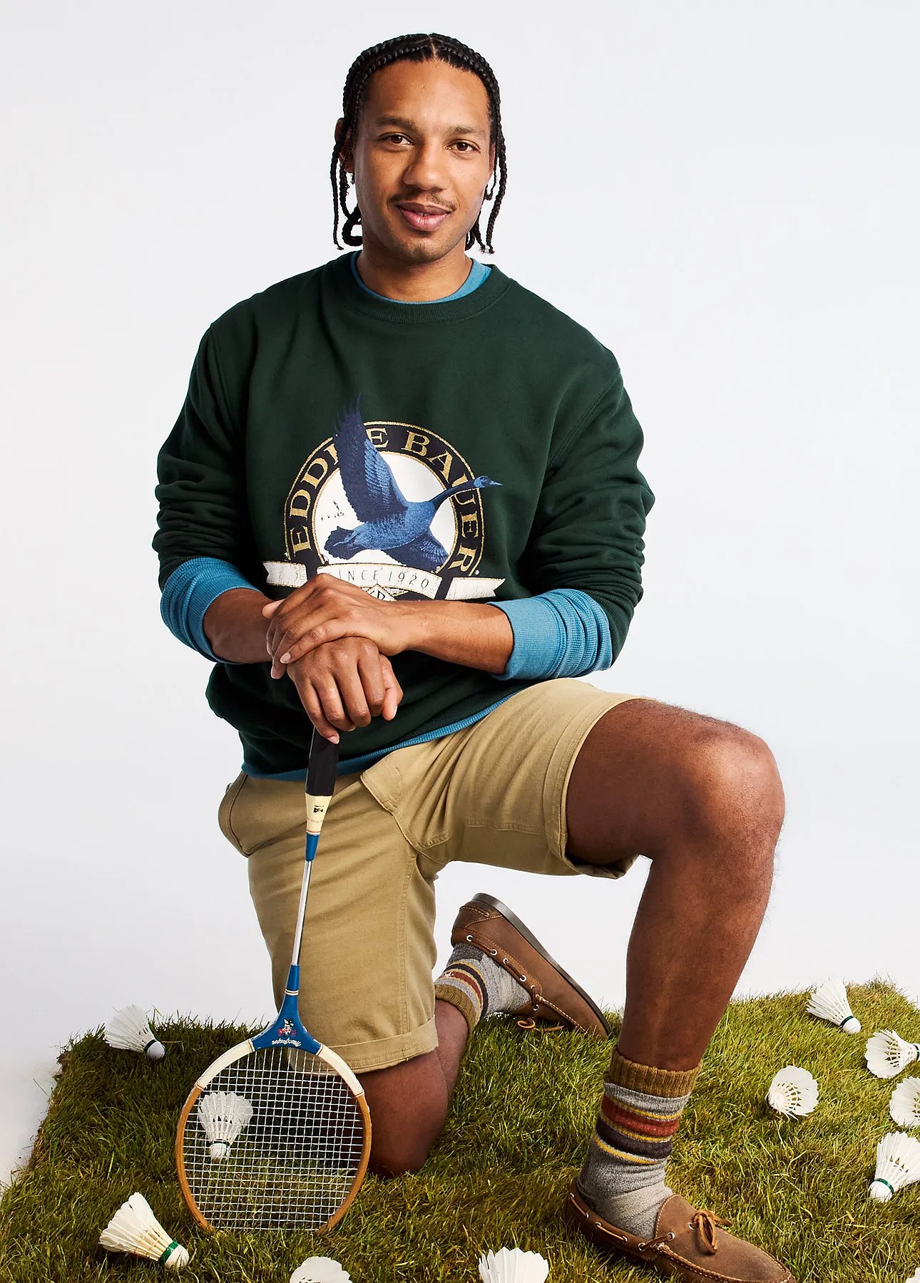 A man wearing a graphic sweatshirt and shorts kneels on the grass and leans on a badminton racket.