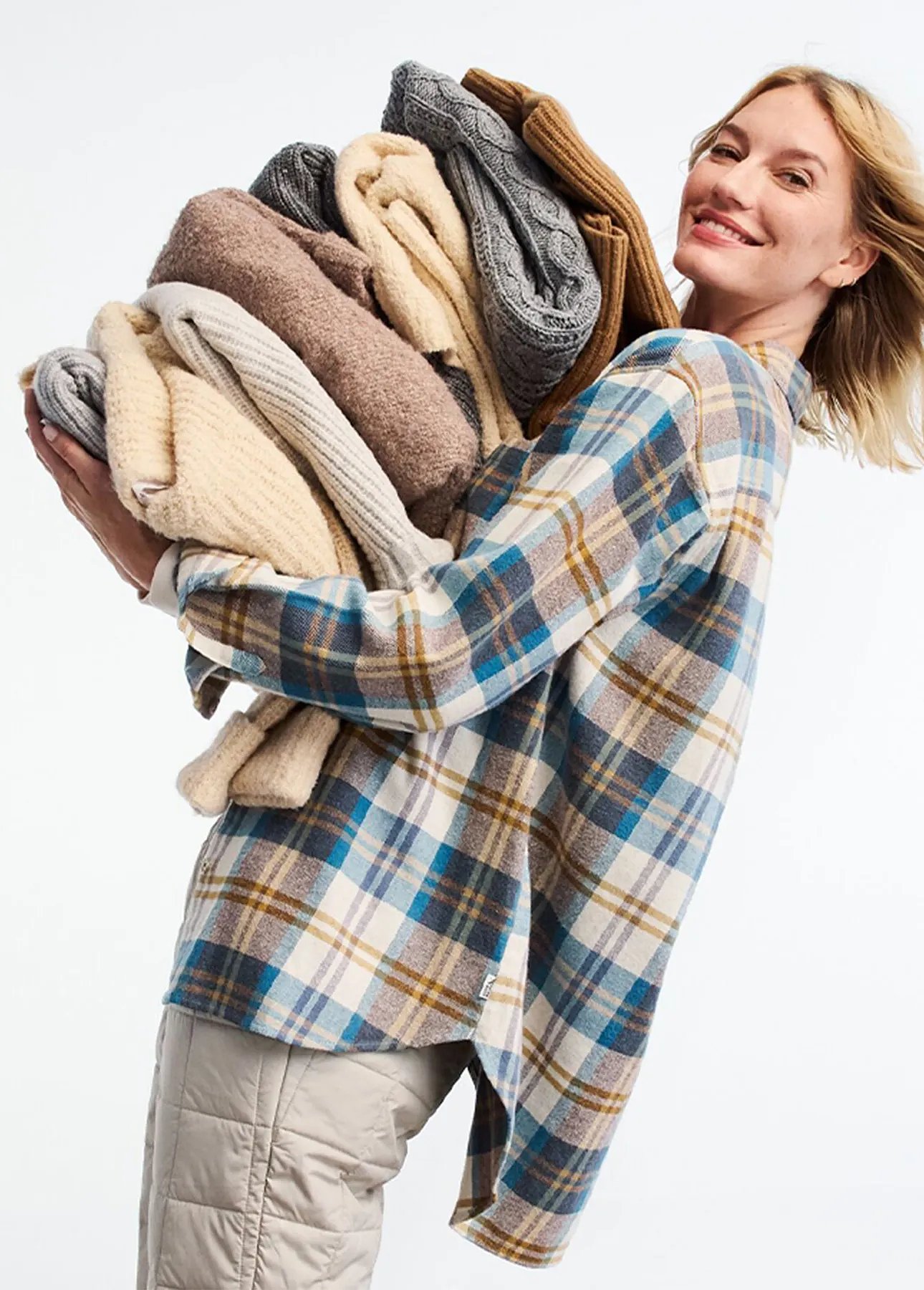 A women wearing a plaid flannel shirt carries a large pile of folded sweater.