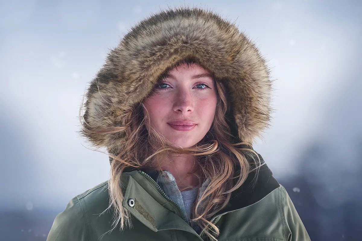 A portrait of a woman wearing a faux fur lined parka with a winter-inspired background
