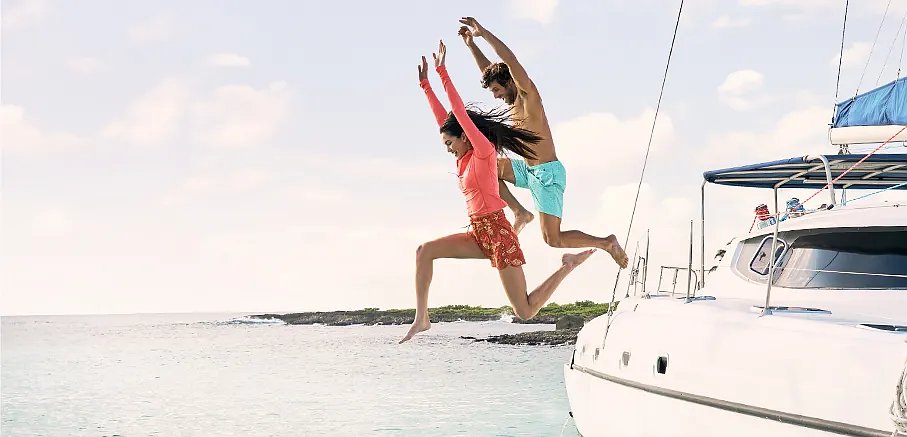 A man wearing swim shorts & a woman wearing a rashguard & swim shorts jump off a boat into the water.
