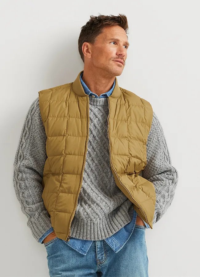 A man wears a grey sweater leaning against a wall.