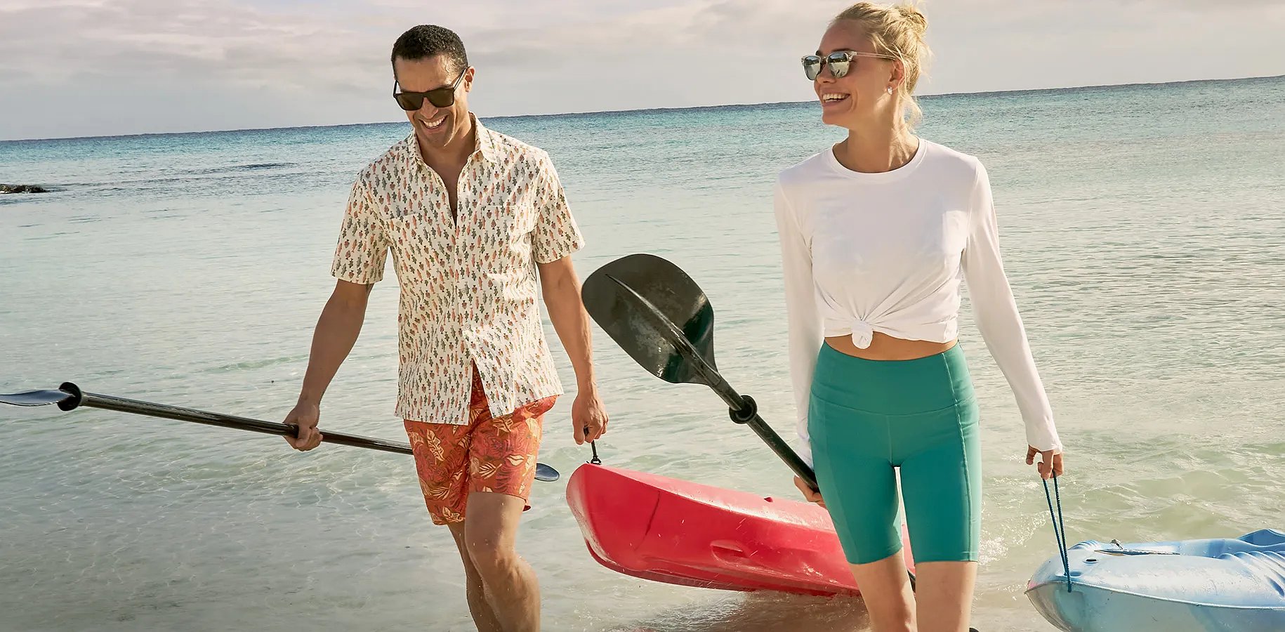 Two people wearing casual shirts carry a kayak and paddles on the beach