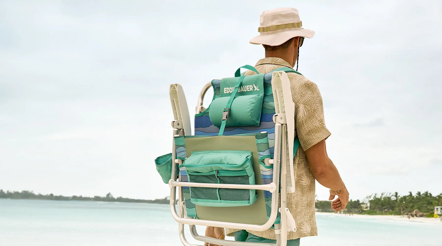A man carrying a backpack chair walks on a tropical beach.