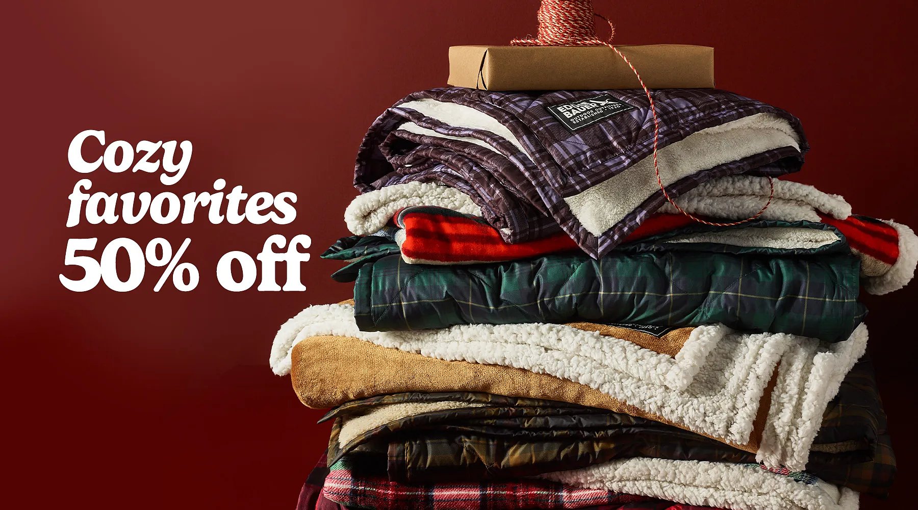50% off Cozy favorites. A stack of folded fleece & down throws with a wrapped gift & a bolt of ribbon on top.