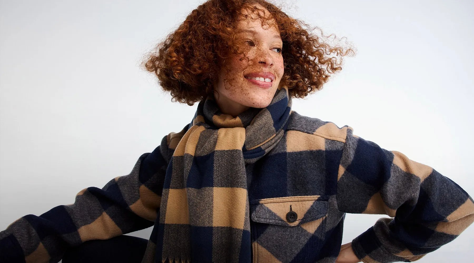 A women with curly hair wears a plaid shirt jacket and a plaid scarf.