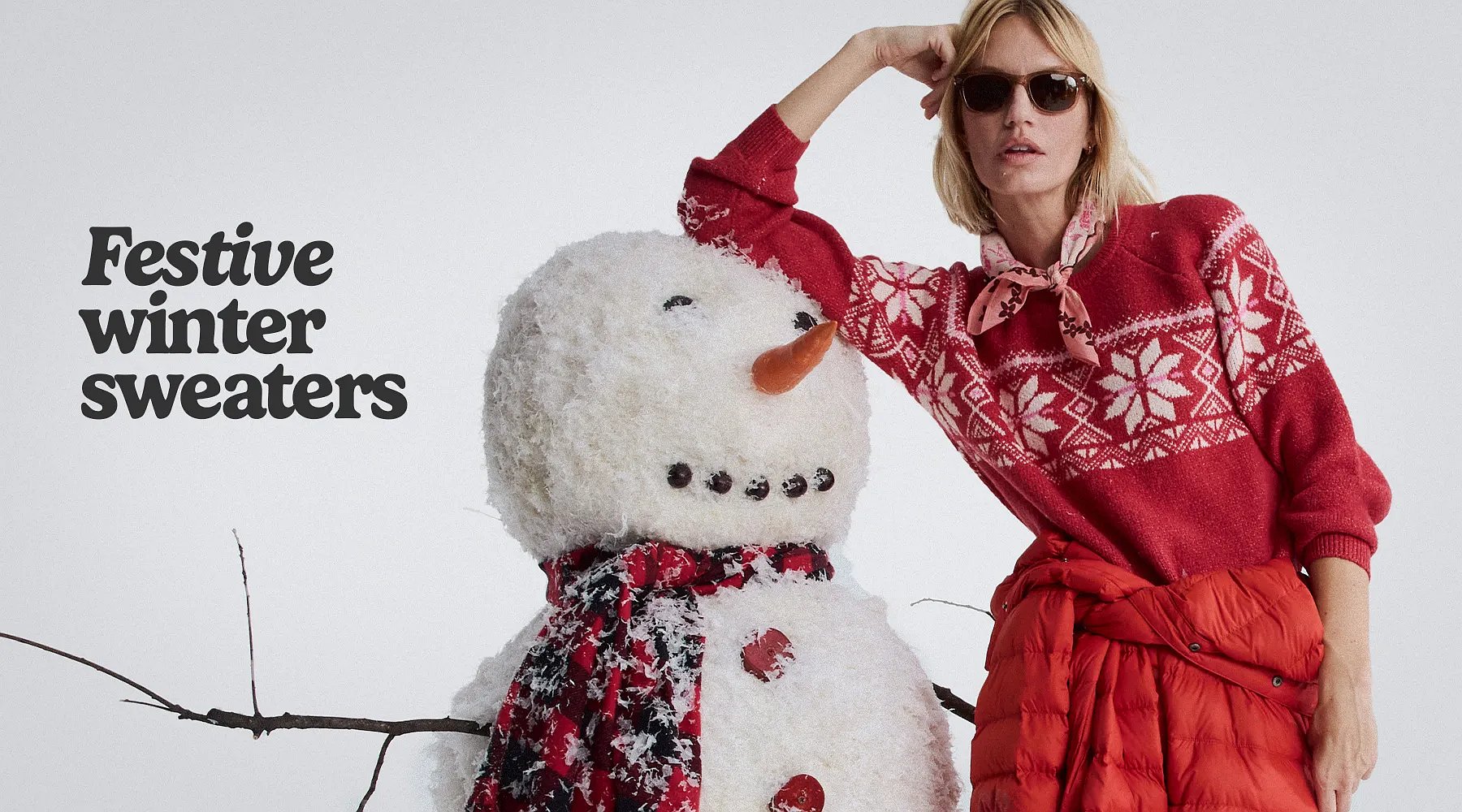 Festive winter sweaters. A women wearing a cozy sweater & down suit tied a the waist leans on against a snowman.