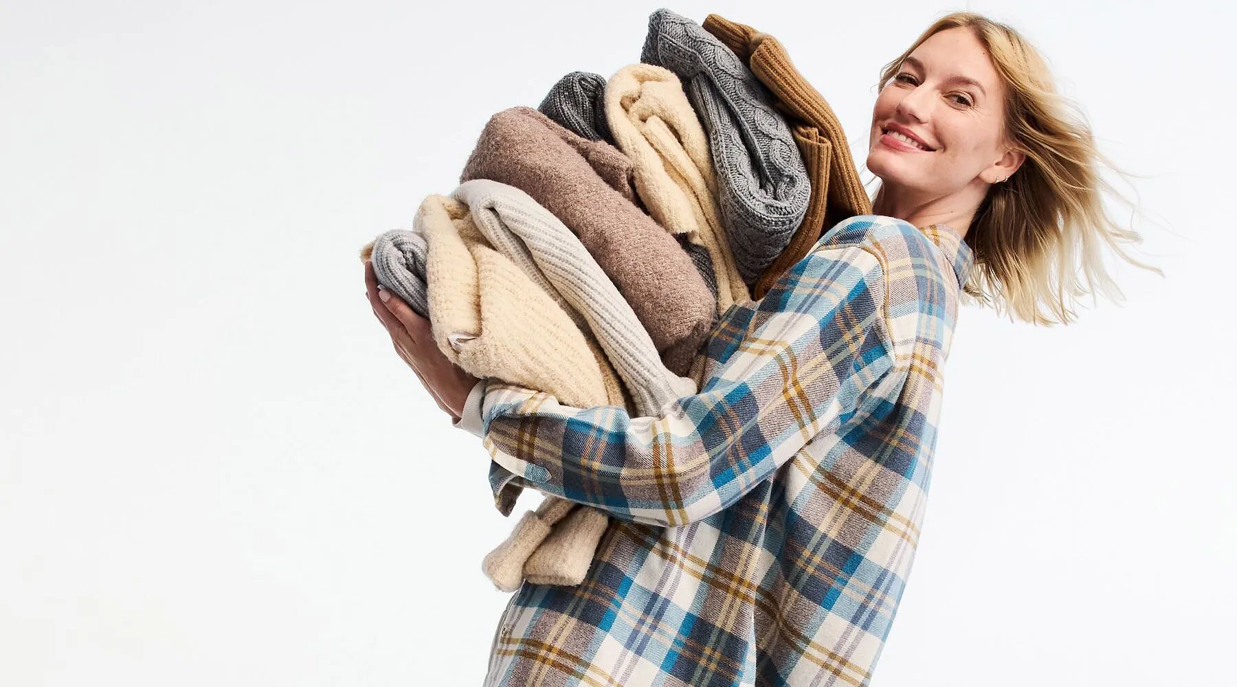 A smiling woman wearing a flannel shirt carries a huge pile of folded sweaters.