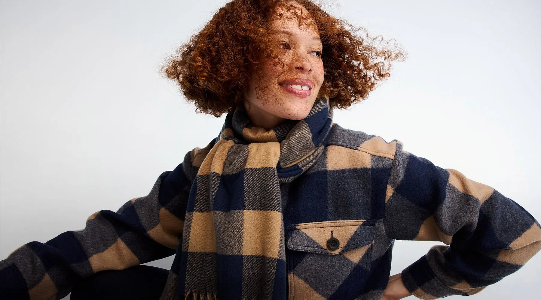 A women with curly hair wears a plaid shirt jacket and a plaid scarf.