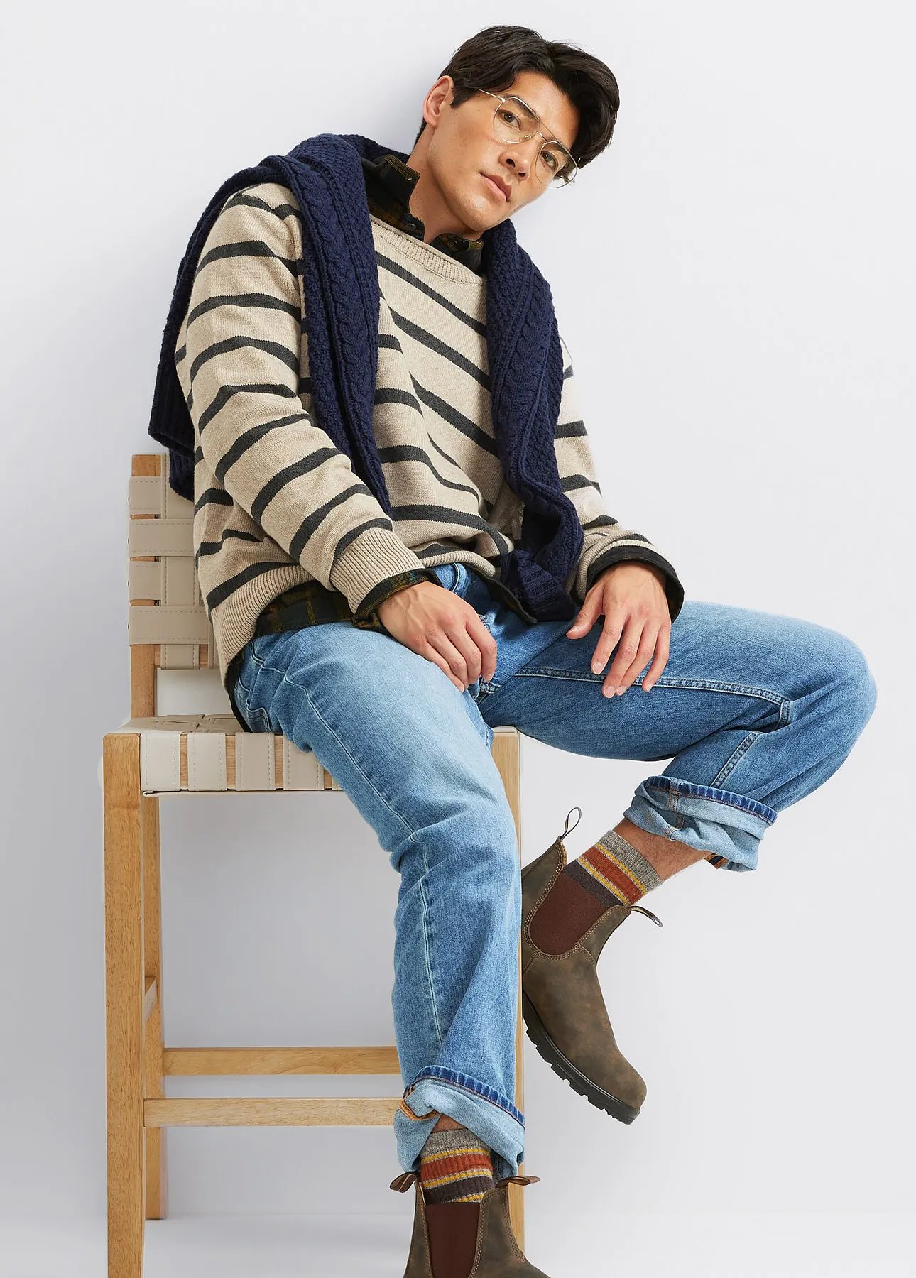 A man wearing jeans, a sweater, and boots sits on a chair.