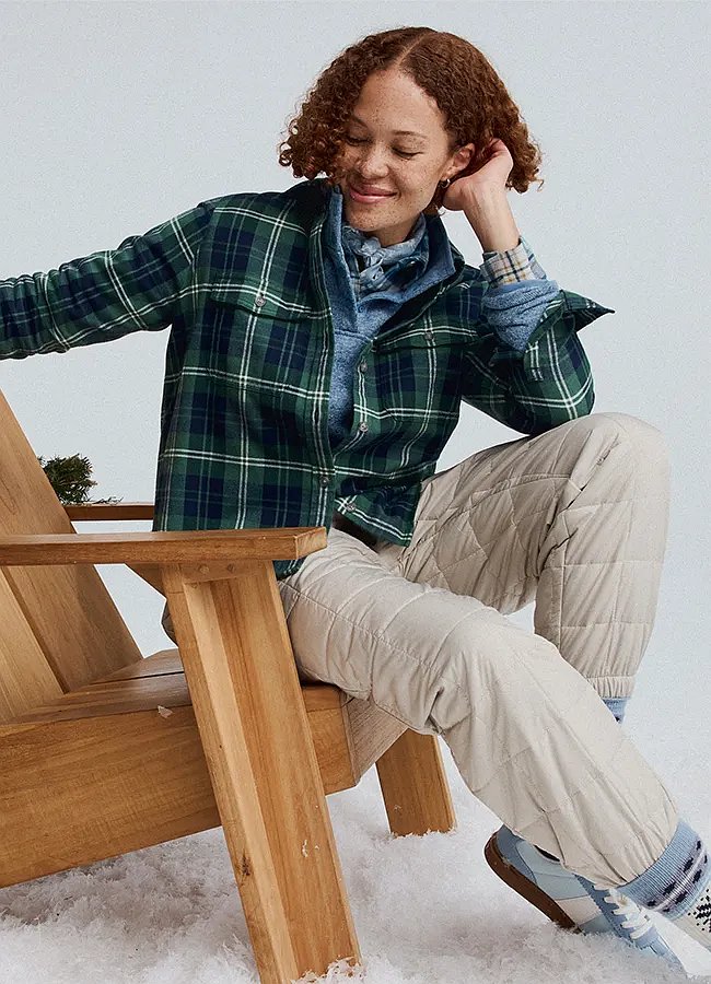 A women wearing a flannel shirt and insulated pants sits in an Adirondak chair.