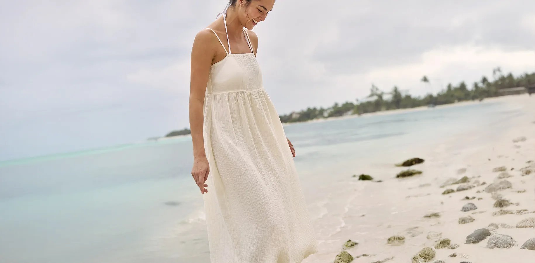 A women wearing a loose, flowing sun dress walks on the beach.