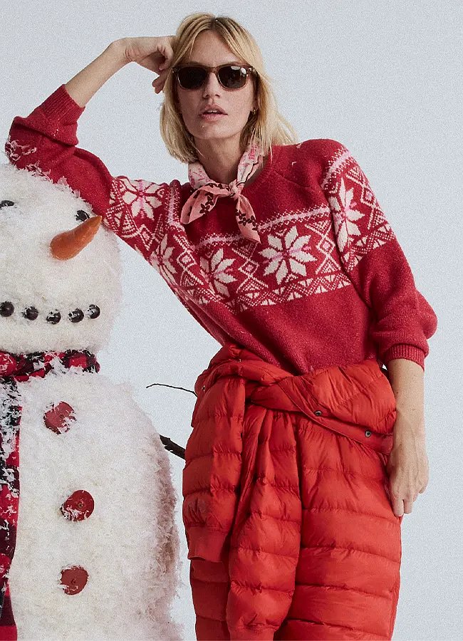 A women wearing a cozy sweater and a down suit tied at the waist leans against a snowman.