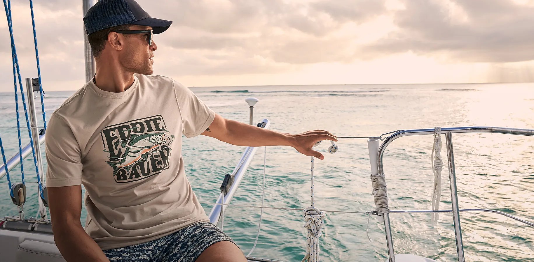 A man in a T-shirt and swim shorts sits on a boat looking out over the water.