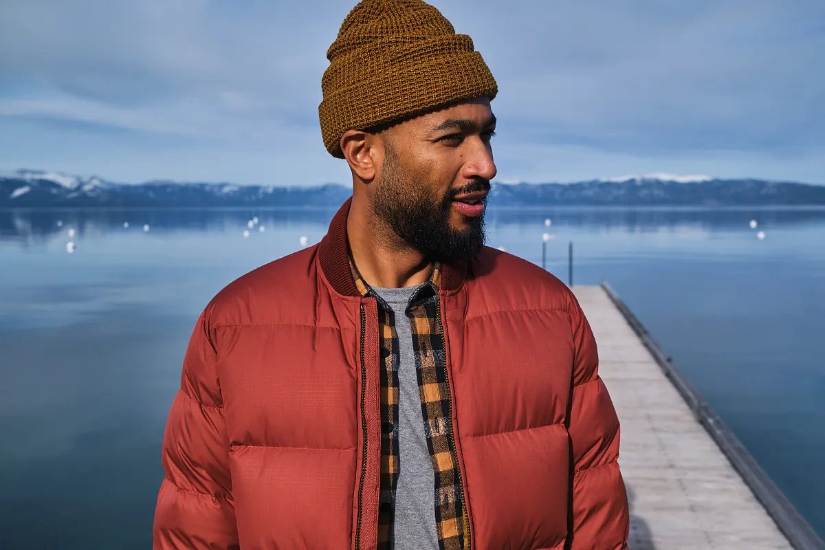 A man wearing a beanie, grey t-shirt, flannel and red insulated jacket standing on dock with a winter-inspired background