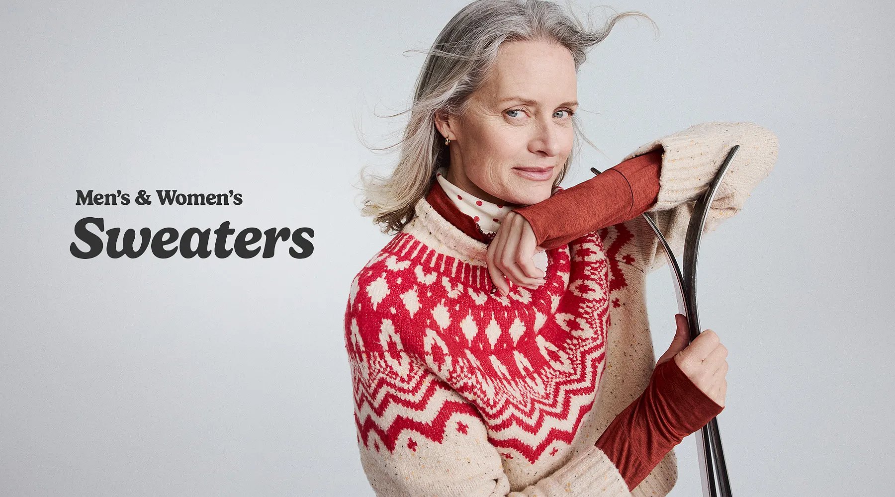 Men’s & women’s sweaters. A woman wearing a fair isle pattern sweater leans on a pair of skies.